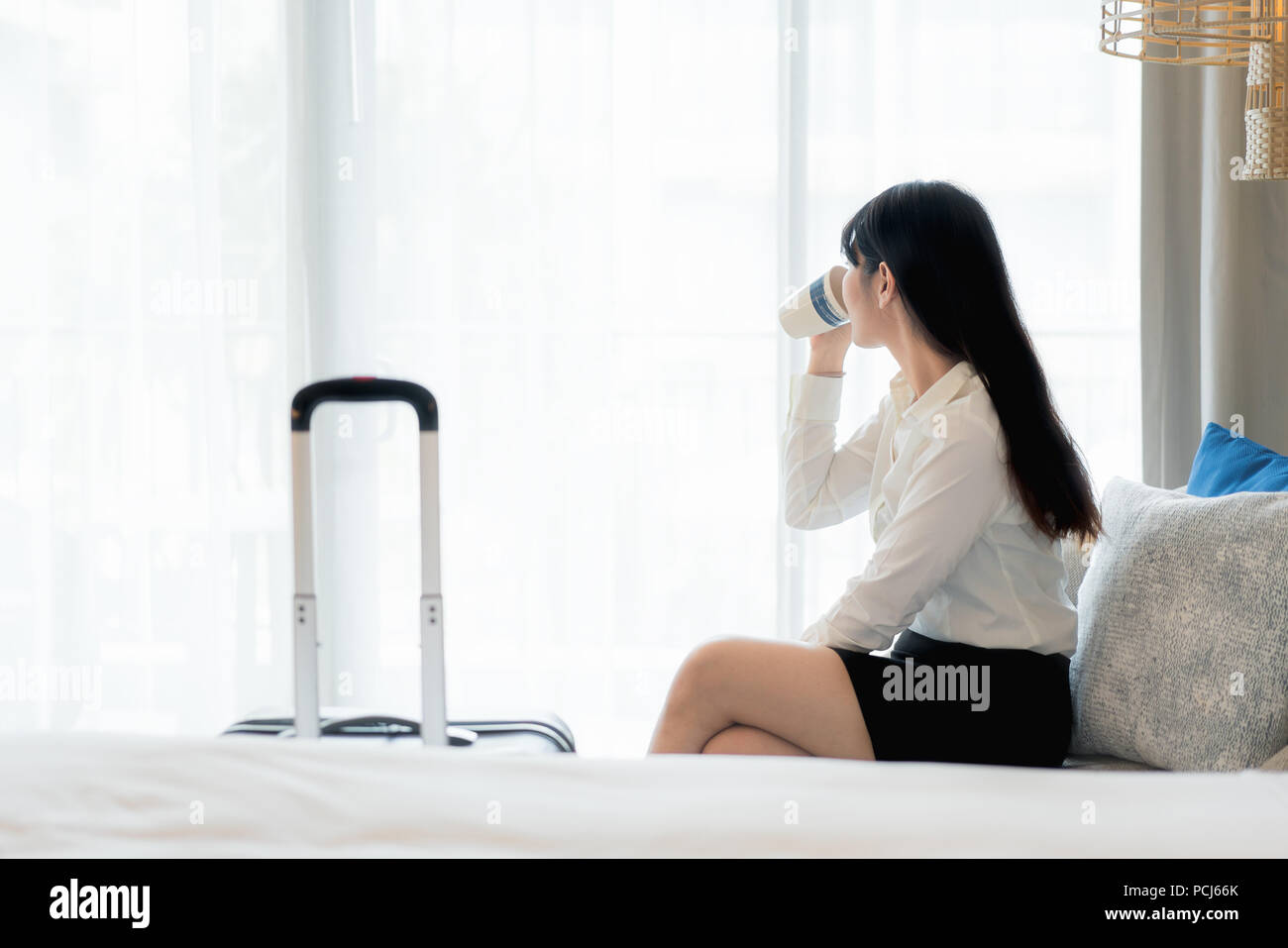 Rilassarsi dopo una dura giornata di lavoro. Bella asiatica giovane imprenditrice sorridente in tuta di bere il caffè e guardare lontano mentre è seduto sul divano in hotel r Foto Stock