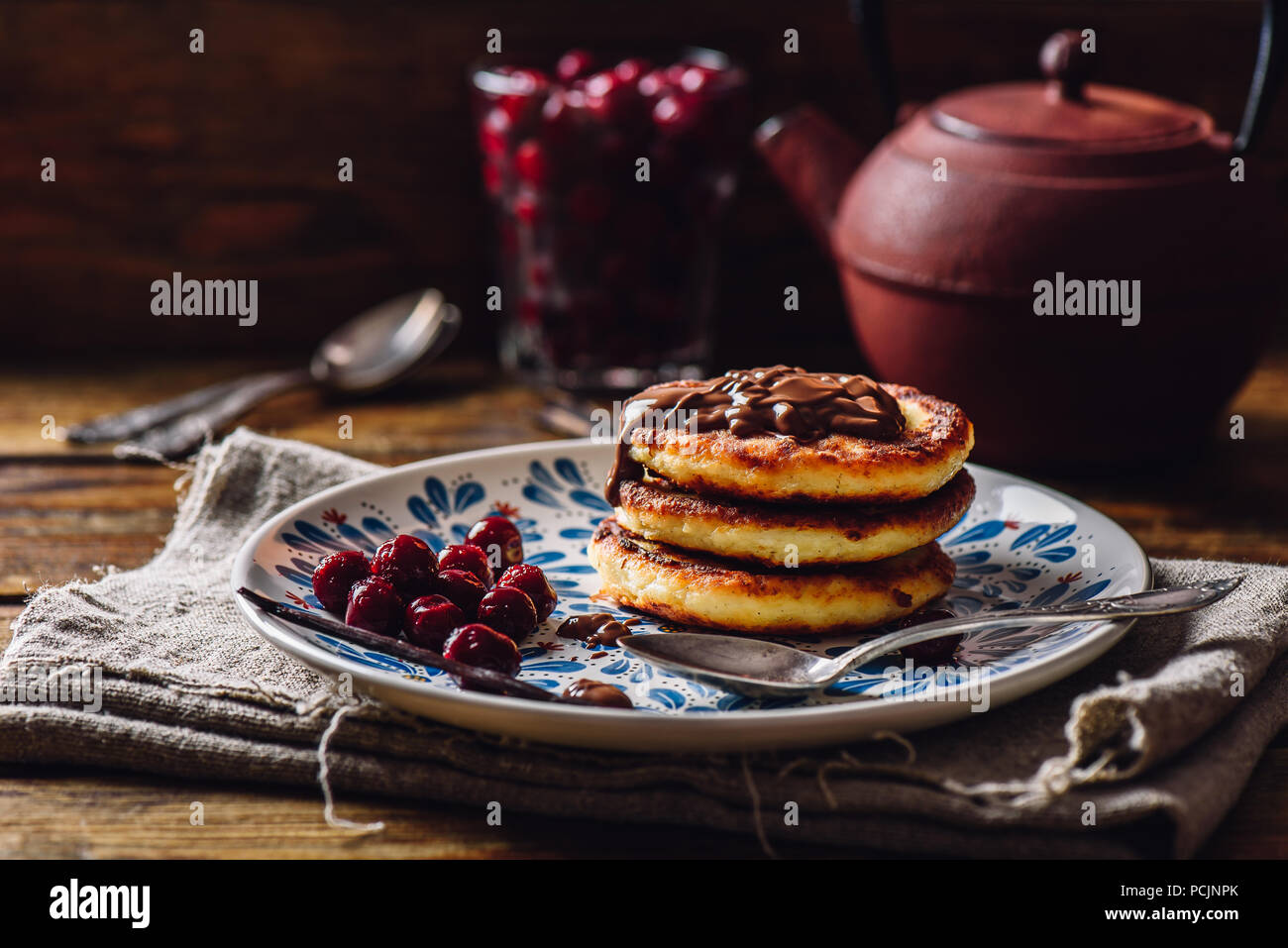 Russo frittelle di Quark con Topping di cioccolato e congelate di ciliegia. Teiera con cucchiai e vetro di bacche sullo sfondo. Foto Stock