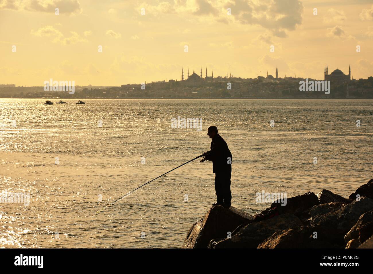 L'uomo è la pesca sulle rocce vicino al faro e la silhouette della città in background Foto Stock