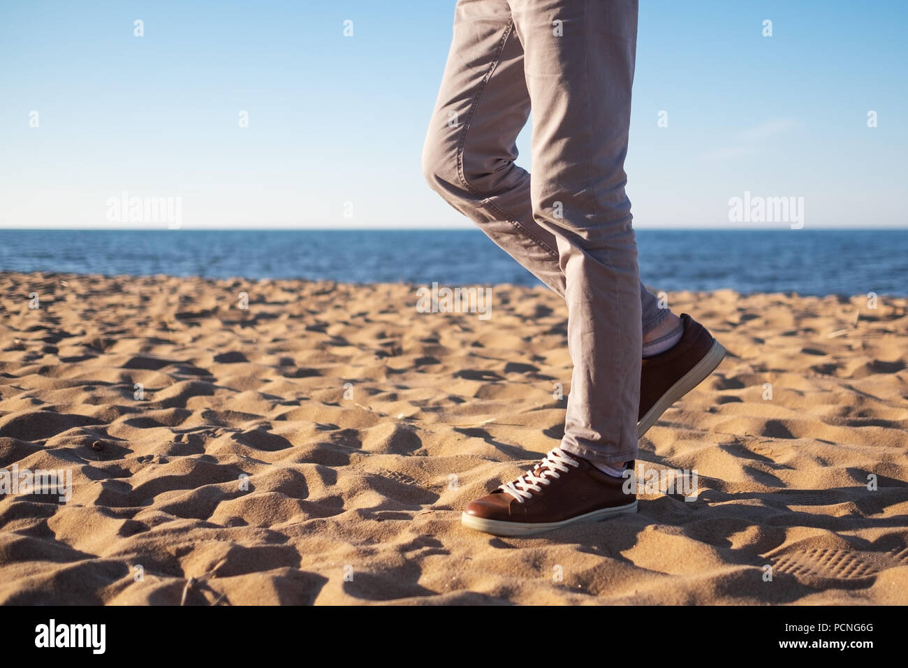 Chiudere l immagine dell uomo gambe camminando da sola spiaggia sabbiosa con oceano blu e bianca sabbia, che indossa pantaloni bianchi e marrone sneakers. Foto Stock
