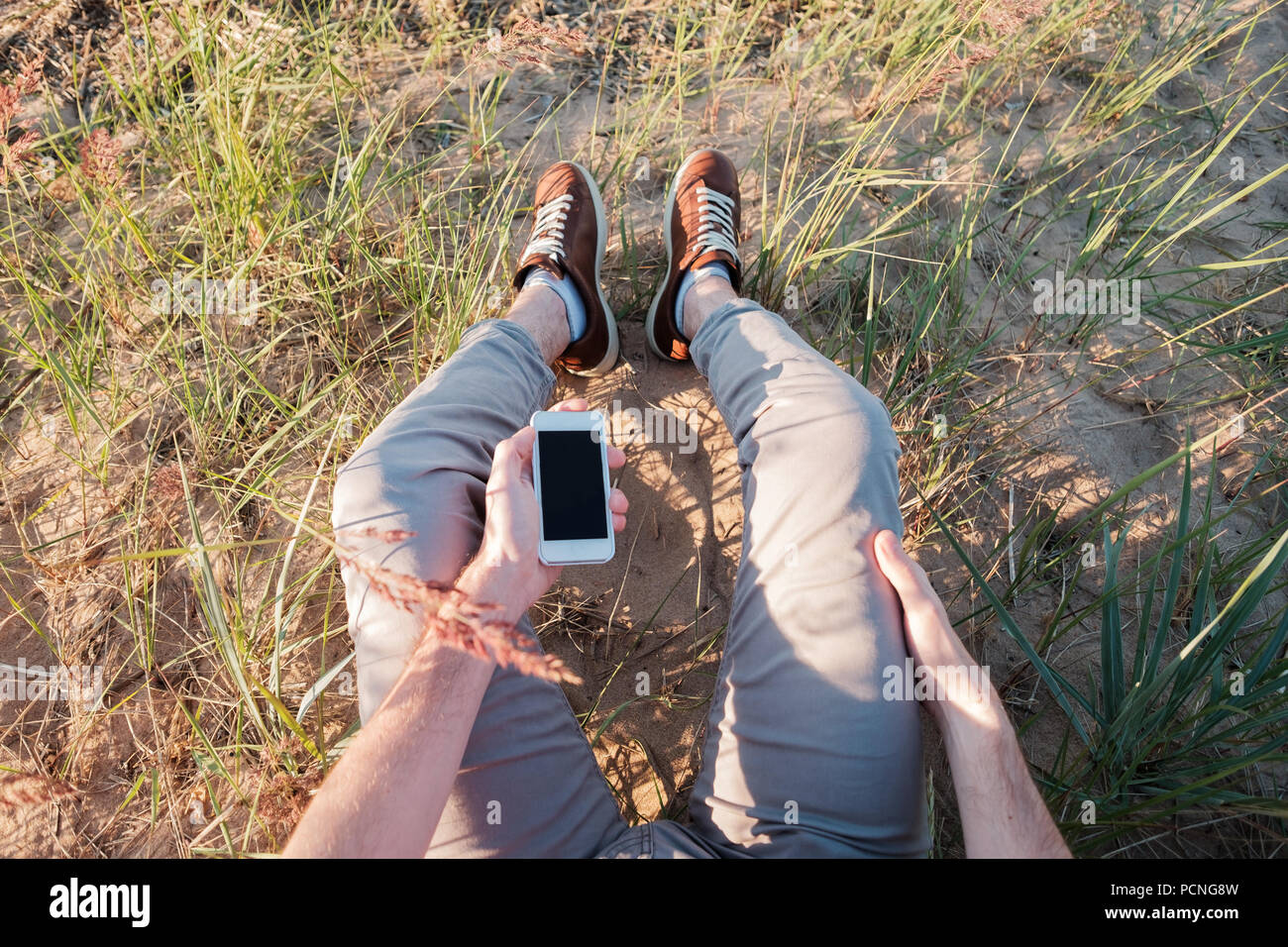 Foto all'aperto dell'uomo seduto sull'erba tenendo il telefono con schermo vuoto. Concetto di essere in contatto in ogni momento in un giorno Foto Stock