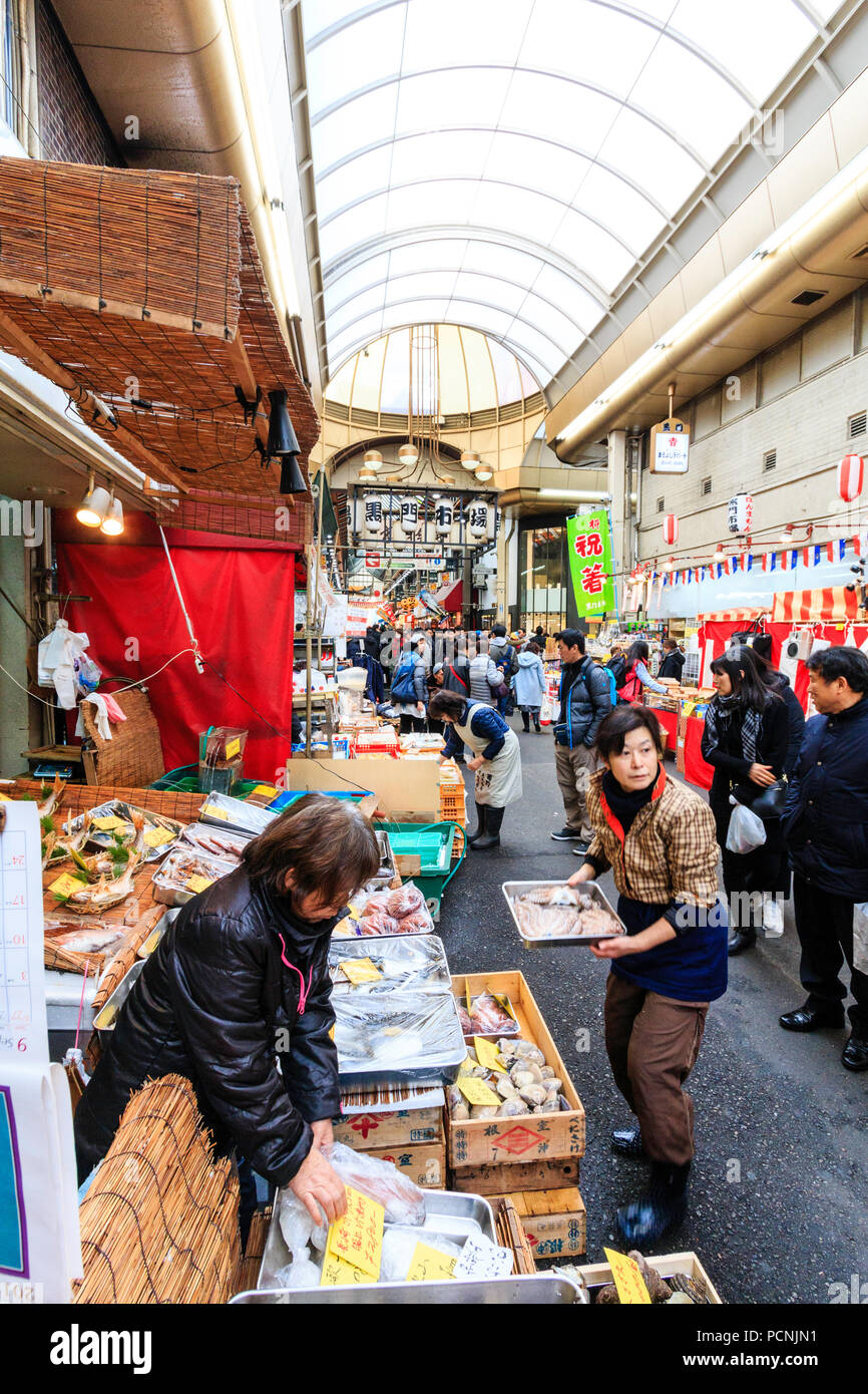 Kuromon Ichiba, mercato alimentare di Osaka. Fish monger stallo con fascio di mare e vongole, due donne assistenti viene visualizzata la regolazione. Arcade affollate di acquirenti Foto Stock