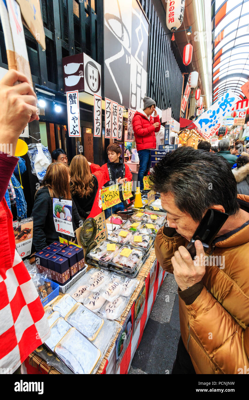 Kuromon Ichiba, mercato alimentare di Osaka. Mercanti di pesce bancarella vendendo preconfezionate seafoods, il cliente in primo piano cercando mentre si utilizza il telefono cellulare. Foto Stock