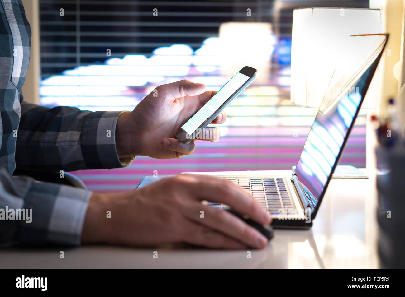 L'uomo utilizza lo smartphone e il computer portatile a tarda notte. Imprenditore o imprenditore nel moderno ufficio di lavoro. Luci della città in background. Foto Stock