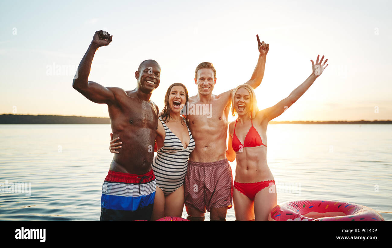 Ridendo gruppo di amici spensierato in piedi a braccetto insieme in un lago divertendosi in un tardo pomeriggio estivo Foto Stock
