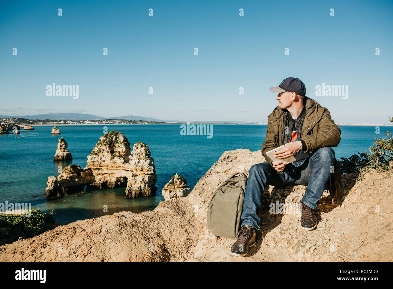 Un giovane uomo o ragazzo o turista utilizza un tablet e guarda lontano nel bellissimo Oceano Atlantico al largo delle coste del Portogallo accanto alla città chiamato Lagos. Foto Stock