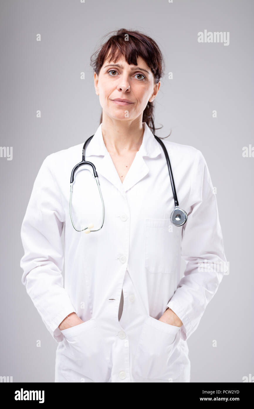 Femmina compassionevole medico o infermiere con uno stetoscopio attorno al collo in piedi con le mani in tasca del suo laboratorio bianco oat cercando simpaticamente Foto Stock