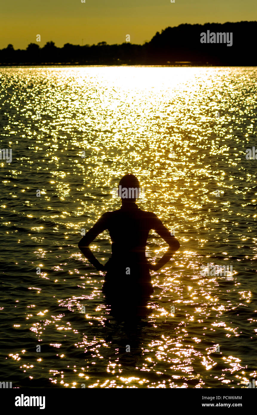 Una donna, mostrato in silhouette, torna alla telecamera, in piedi nel lago Maxinkuckee di Culver, Indiana, indossando una muta e nuotare cap guardando ad ovest verso il sole di setting Foto Stock