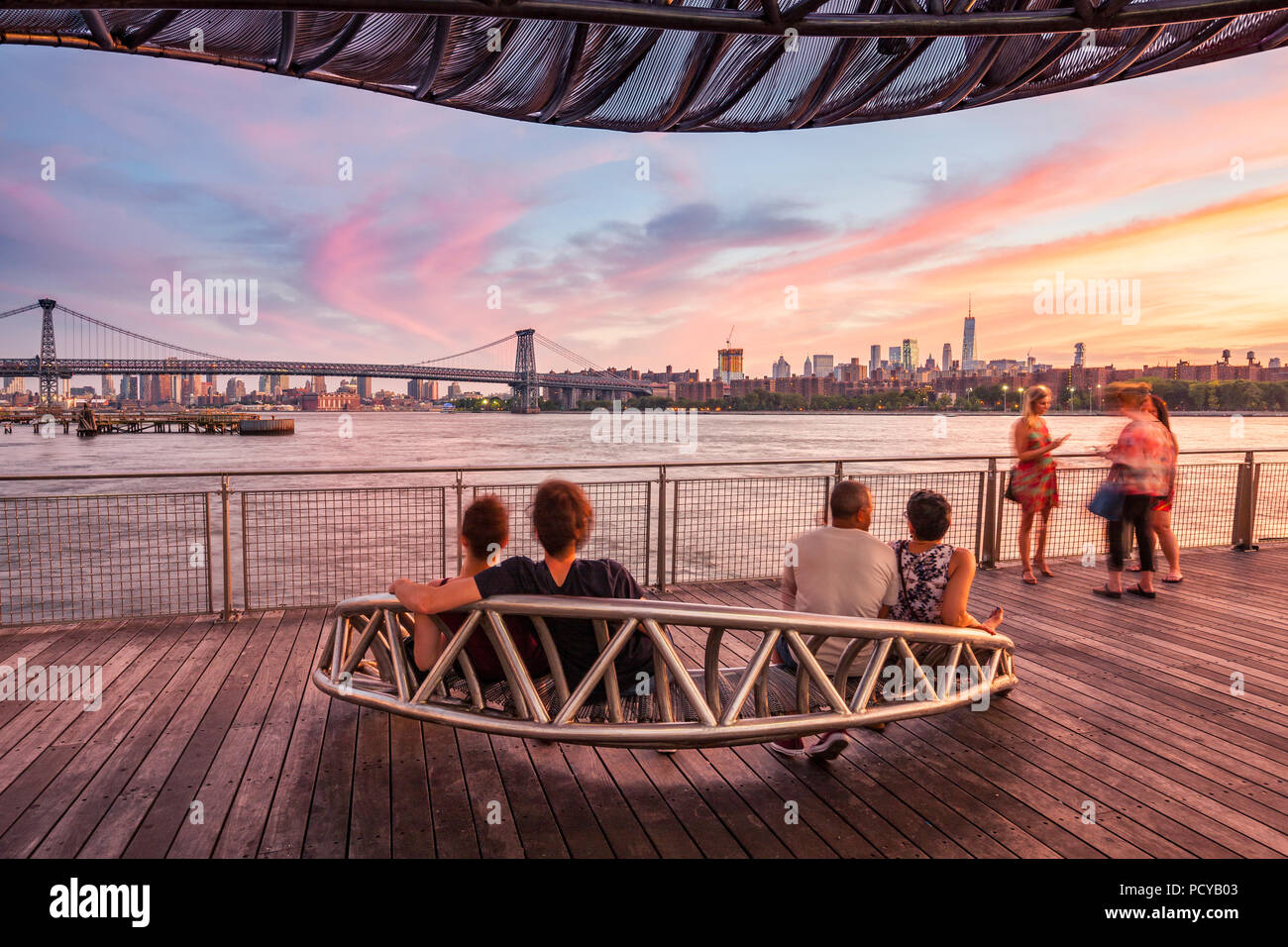 La Gente seduta sul banco di lavoro e godendo la vista a Williamsburg Bridge a le nuvole colorate al tramonto Foto Stock