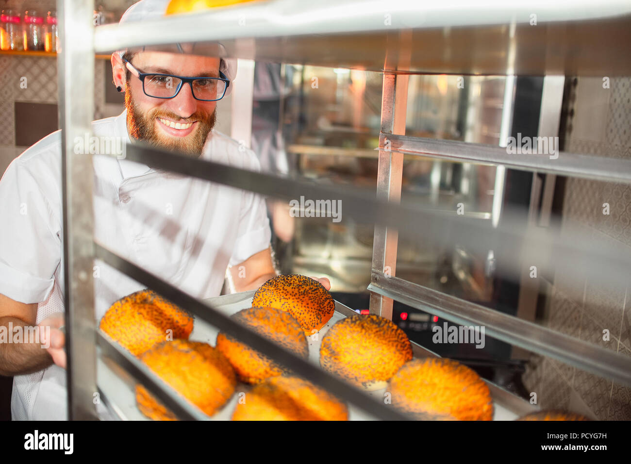 Un giovane bello baker azienda panini freschi con semi di papavero in background del forno. Foto Stock