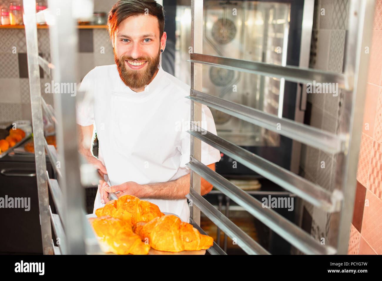 Un giovane bello baker prende freschi croissants caldi dal forno su una pala. Foto Stock