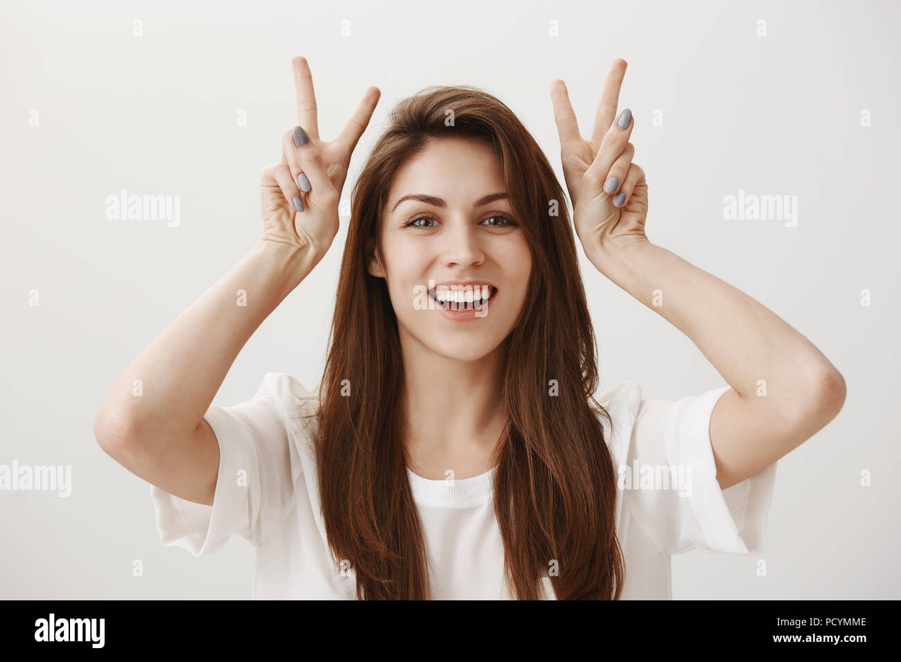 Affascinante ragazza rendendo l'aria preventivi. Ritratto di affascinante giovane femmina alzando le mani in prossimità della testa e mostra v o segno di pace, sorridente largamente, dicendo ferro da stiro Foto Stock