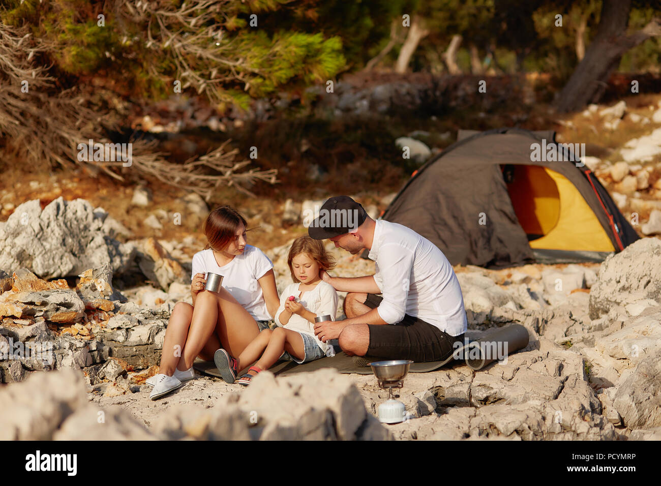 Gentile famiglia giovane, seduto sul pad di terra vicino alla tenda sul litorale di pietra di giorno. Papà nel cappuccio, versando il tè da thermos in tazza di detenzione da parte sua litt Foto Stock