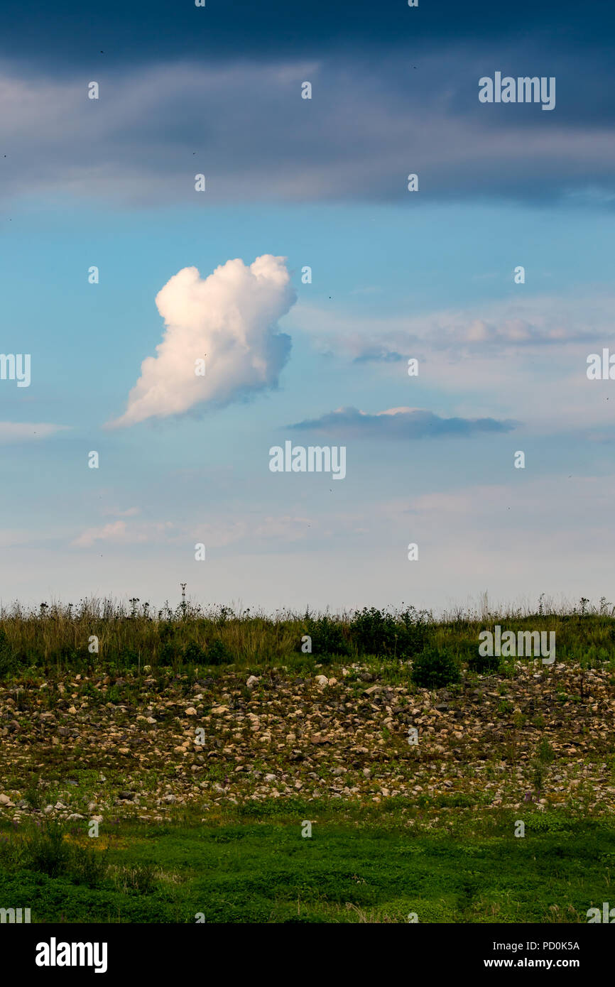 A forma di fantasma nuvola nel cielo blu, ciottoli ed erba nella parte anteriore e tranquillo pomeriggio estivo paesaggio verticale con colorati e nuvole contrasty Foto Stock