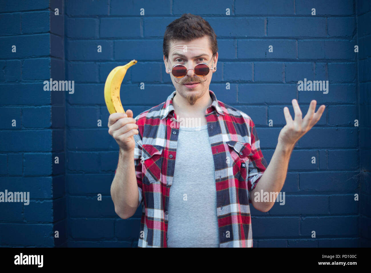 Giovane uomo bianco in hipster vestiti, con parentesi di baffi e occhiali vintage, tenendo una banana in una mano in piedi di fronte ad un blu un muro di mattoni. Foto Stock