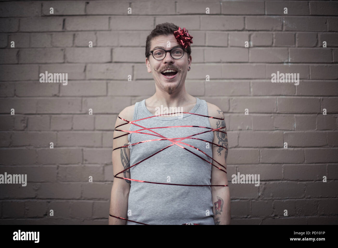 Giovane uomo bianco con tatuaggi avvolto in un regalo rossa nastro con un  fiocco rosso nel lato della testa, ricci di baffi e occhiali vintage Foto  stock - Alamy
