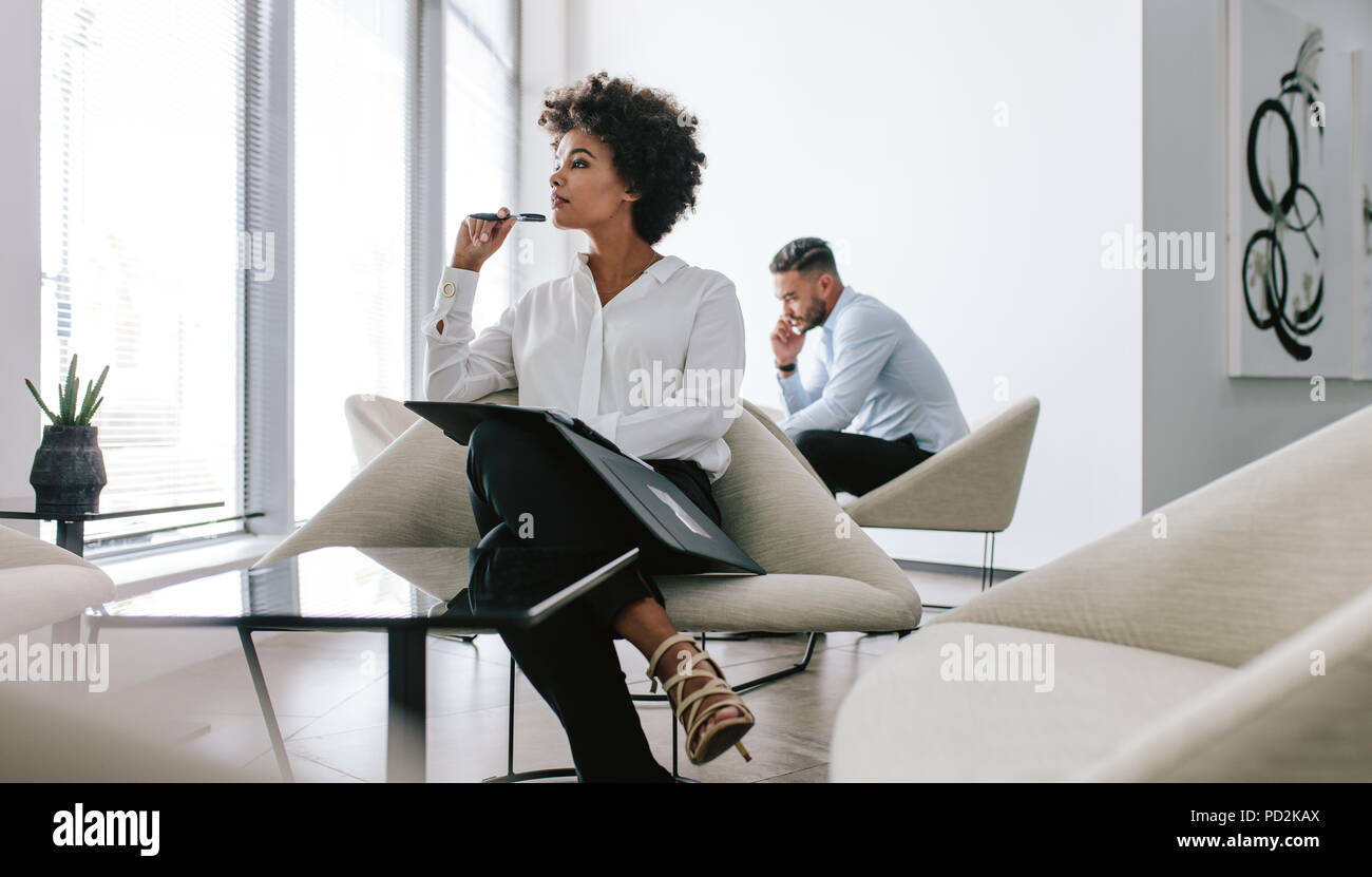 Giovane donna seduta nel corridoio di office con un file e guardando lontano pensando. Business donna riflettendo su una nuova proposta commerciale con un collega che parla Foto Stock