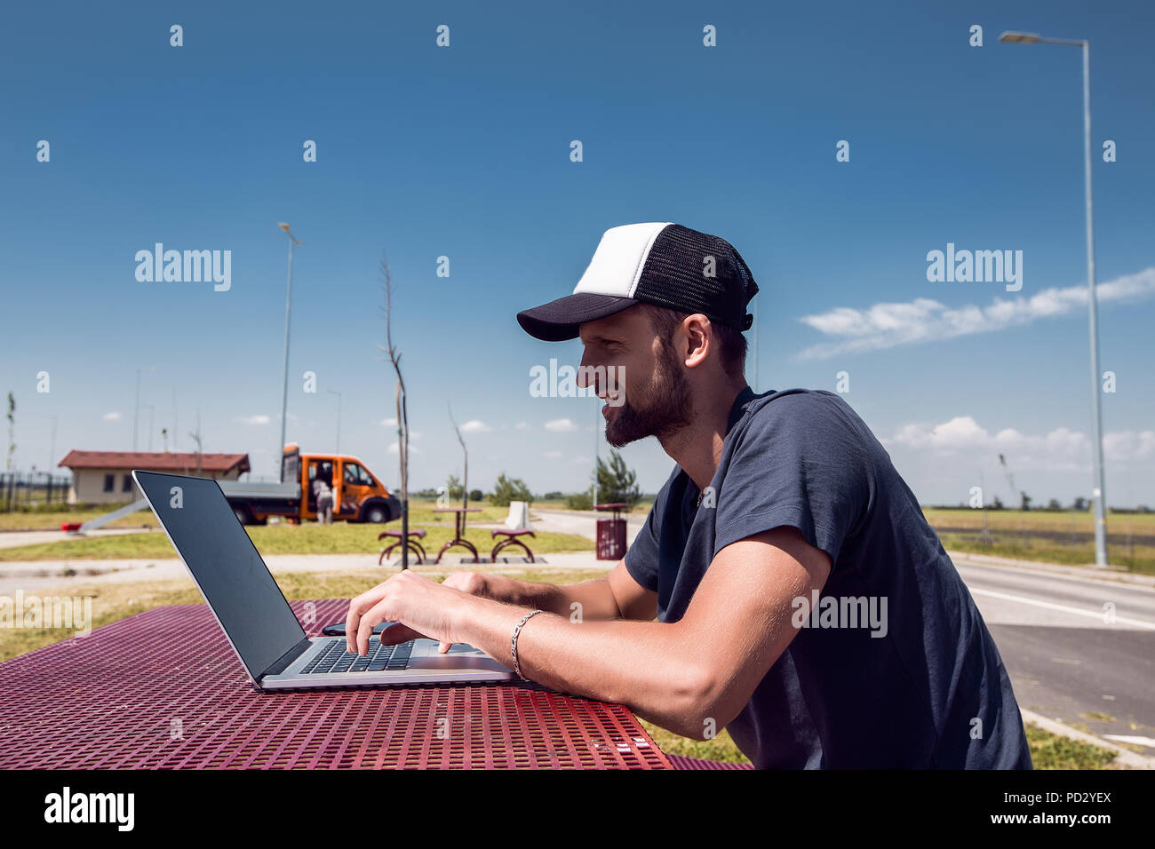 Uomo in cappello da baseball seduti al picnic sul ciglio della strada tabella digitando su laptop Foto Stock