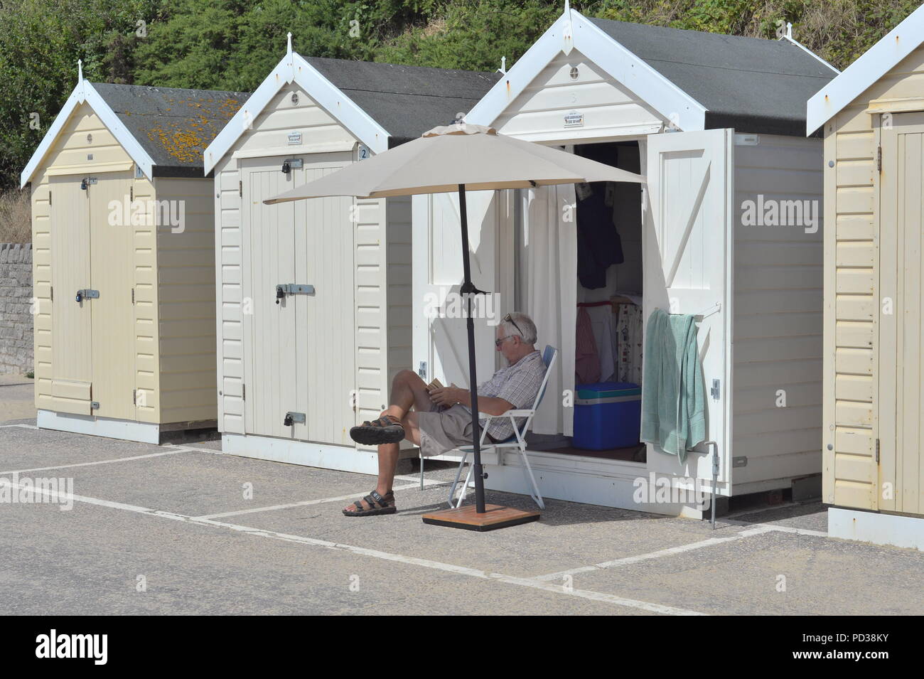 Uomo seduto su una sedia che legge fuori da una capanna sulla spiaggia. Bournemouth, Dorset, Regno Unito, lunedì 6th agosto 2018. Il tempo caldo sulla spiaggia della costa meridionale mentre la gloriosa ondata di caldo estiva continua. Foto Stock