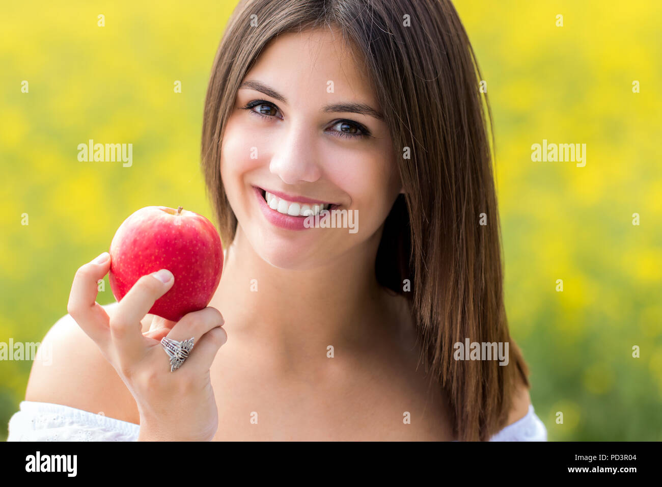 Close up faccia colpo di attraente giovane donna con sorriso affascinante e sorriso toothy azienda red apple contro il fiore giallo dello sfondo. Foto Stock