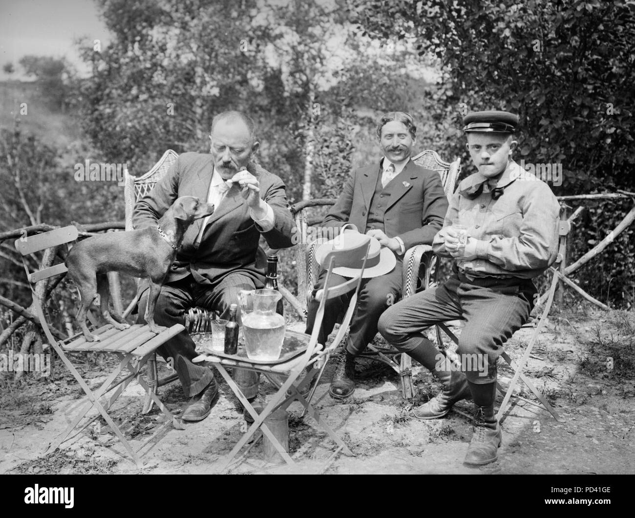 Un trio di uomini godetevi un drink al di fuori con il cane, ca. 1910. Foto Stock