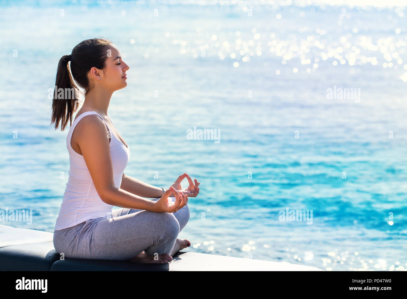 A piena lunghezza Ritratto di giovane donna meditando accanto al mare. Vista laterale della ragazza facendo esercizi yoga in inizio di mattina di sole con riflesso luminoso sull'acqua Foto Stock