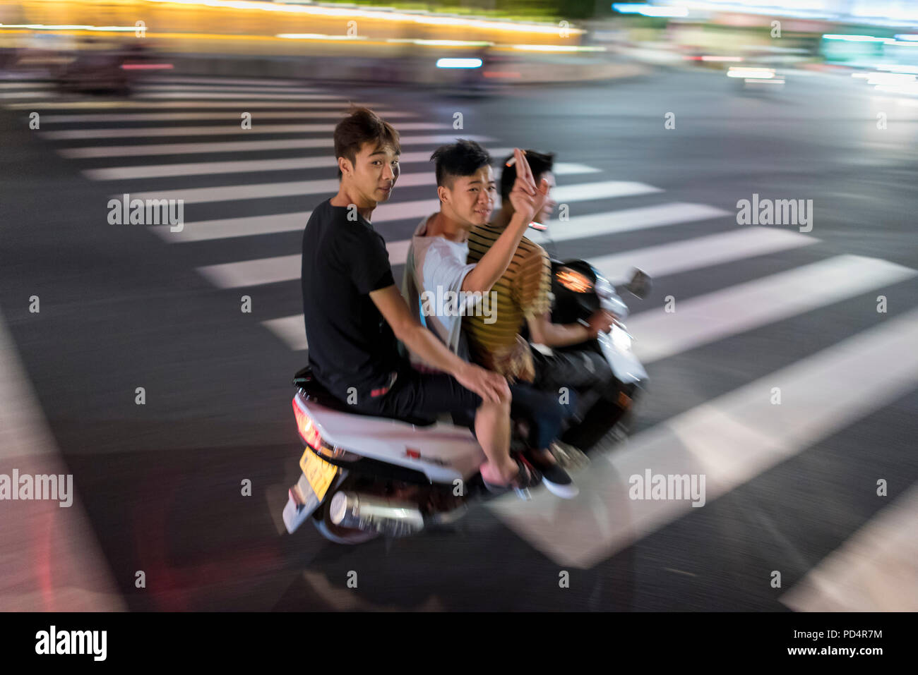 Tre ragazzi teen wave mentre passano su scooter velocità attraverso il centro cittã sulla calda notte d'estate, Libo, Guizhou, Cina. Foto Stock