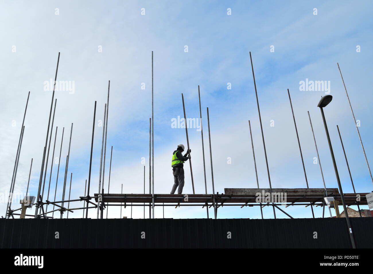 Silhouette di un lavoratore edile azienda tagliere in legno sulla parte superiore del nuovo sviluppo circondato bay pali metallici contro il cielo blu Foto Stock