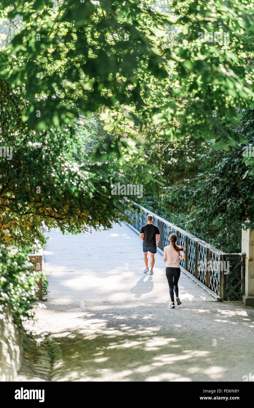 Coppia giovane camminando lungo il tranquillo paesaggio del parco in una bella giornata d'estate. Baldacchino verde di alberi e di un ponte metallico sopra l'acqua di uno stagno. Foto Stock