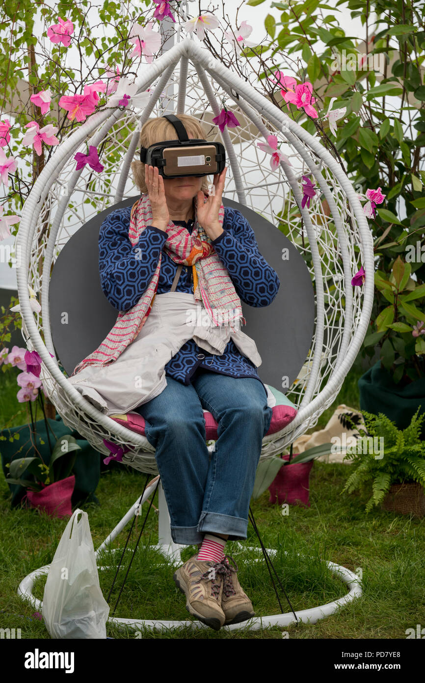 Donna che indossa le cuffie VR, seduto al posto di guida tra i fiori in realtà virtuale esperienza di orchidee - RHS Chatsworth Flower Show, Derbyshire, Inghilterra, Regno Unito. Foto Stock