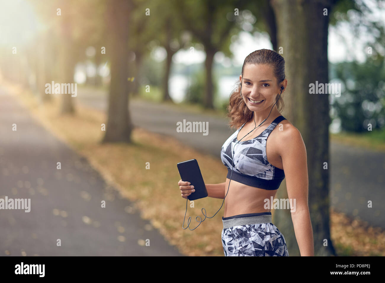 Attraente donna sportivo ascolta musica sul suo telefono cellulare jogging su una strada alberata passando al sorriso felicemente con la videocamera Foto Stock