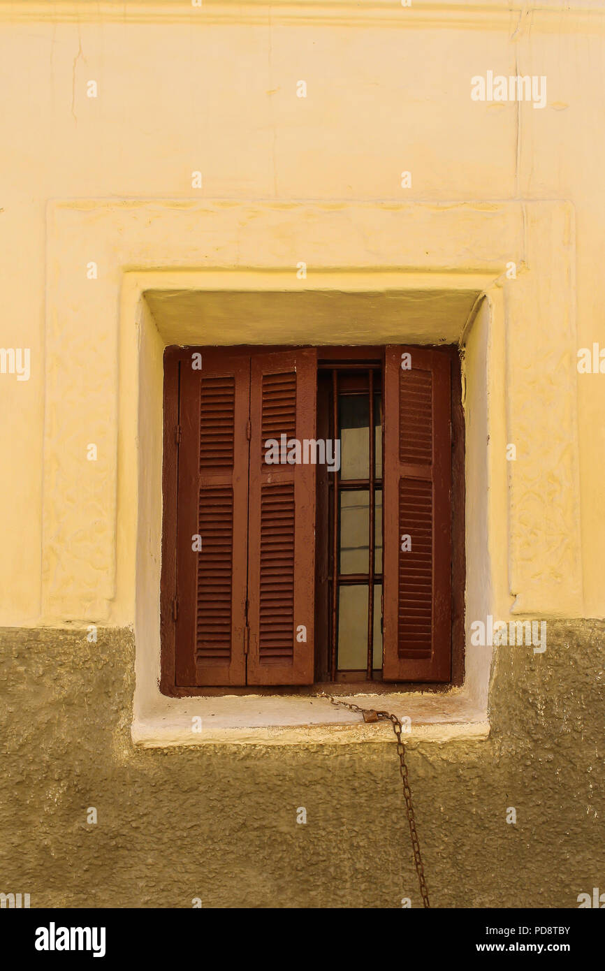 Il giallo muro di una casa con una finestra con apertura parziale otturazione marrone. Catena arrugginita appeso alla finestra. El Jadida, Marocco. Foto Stock