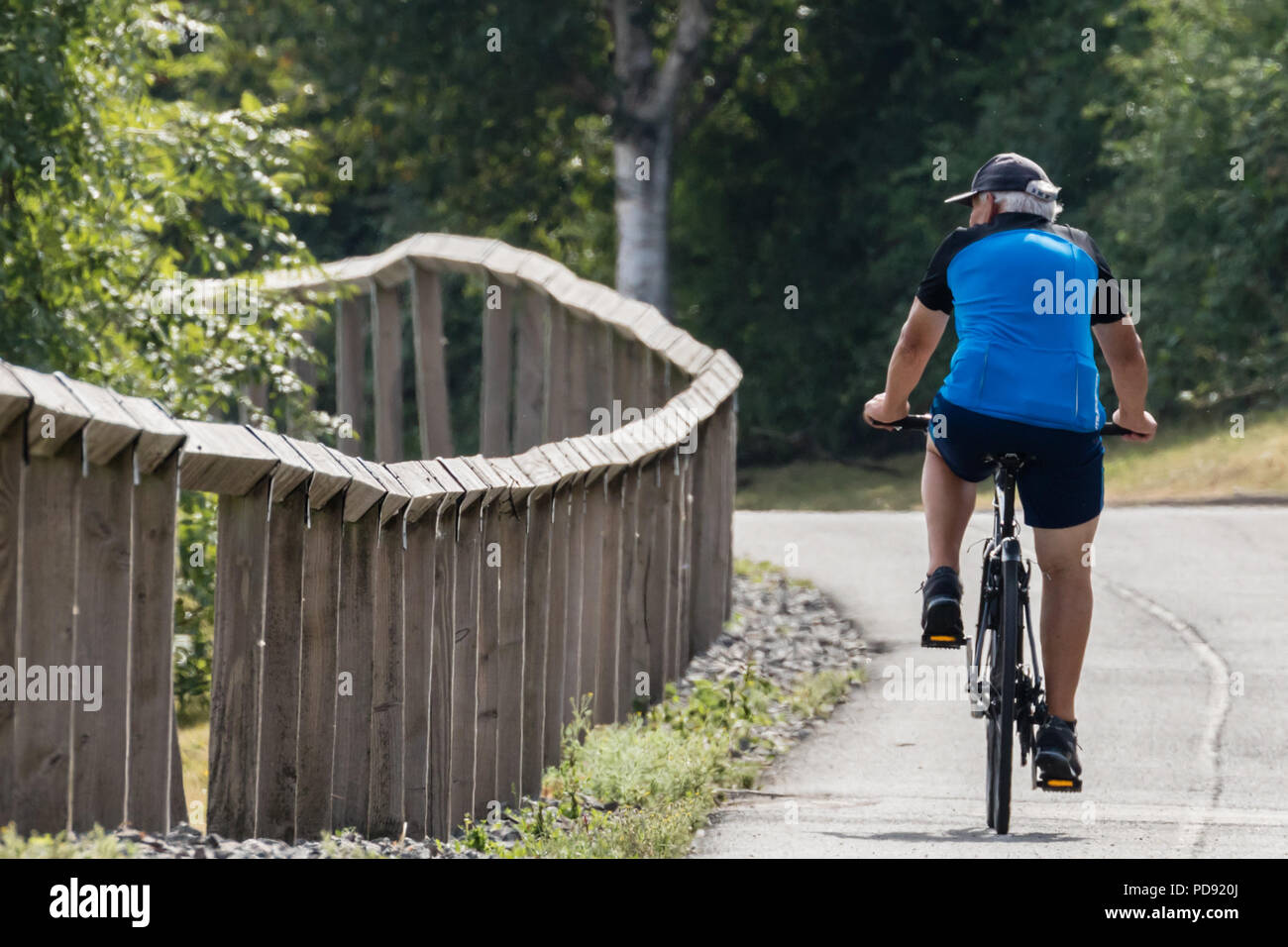 Draycote acqua, 07-09-18. Anziani ciclista indossando un cappello pedali la sua bicicletta su una strada a fianco di una recinzione che le curve a sinistra e al di fuori della vista. Foto Stock