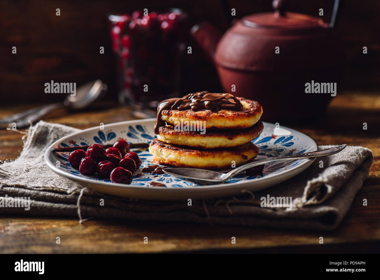 La prima colazione con frittelle di Quark con pasta cioccolato e congelate di ciliegia. Teiera con cucchiai e vetro di bacche sullo sfondo. Foto Stock