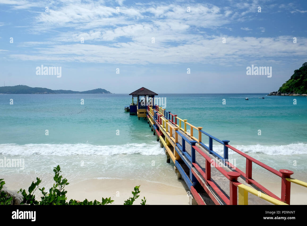 Calcestruzzo colorato e pontile in legno sporgente su una spiaggia di sabbia bianca di un blu limpido mare calmo, rocce e alberi su entrambi i lati il cielo è blu con whi Foto Stock