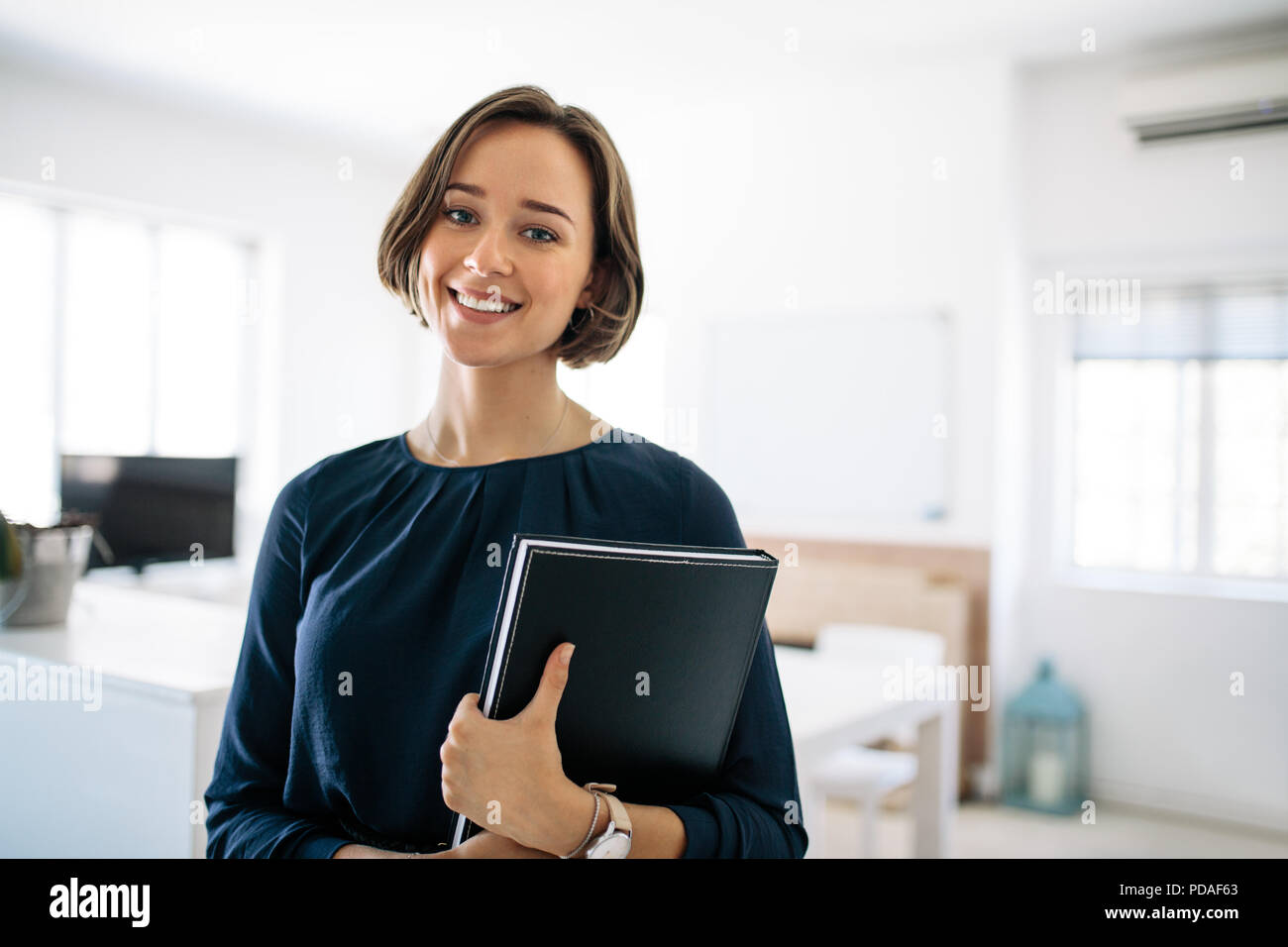 Imprenditrice sorridente in piedi in ufficio tenendo un diario. Ritratto di una donna felice imprenditore al lavoro. Foto Stock