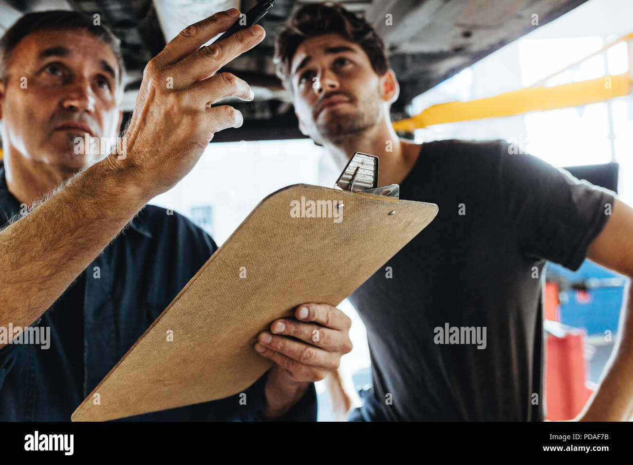 Due meccanici esperti in piedi sotto un auto e ispezionando il problema. Meccanica facendo auto ispezione con appunti. Foto Stock