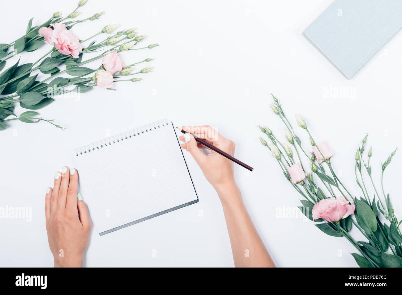 Donna con la mano una matita rendendo note sul foglio bianco vicino a libro e bouquet di fiori, vista dall'alto. Piano di composizione laico femminile stilizzata sul posto di lavoro. Foto Stock