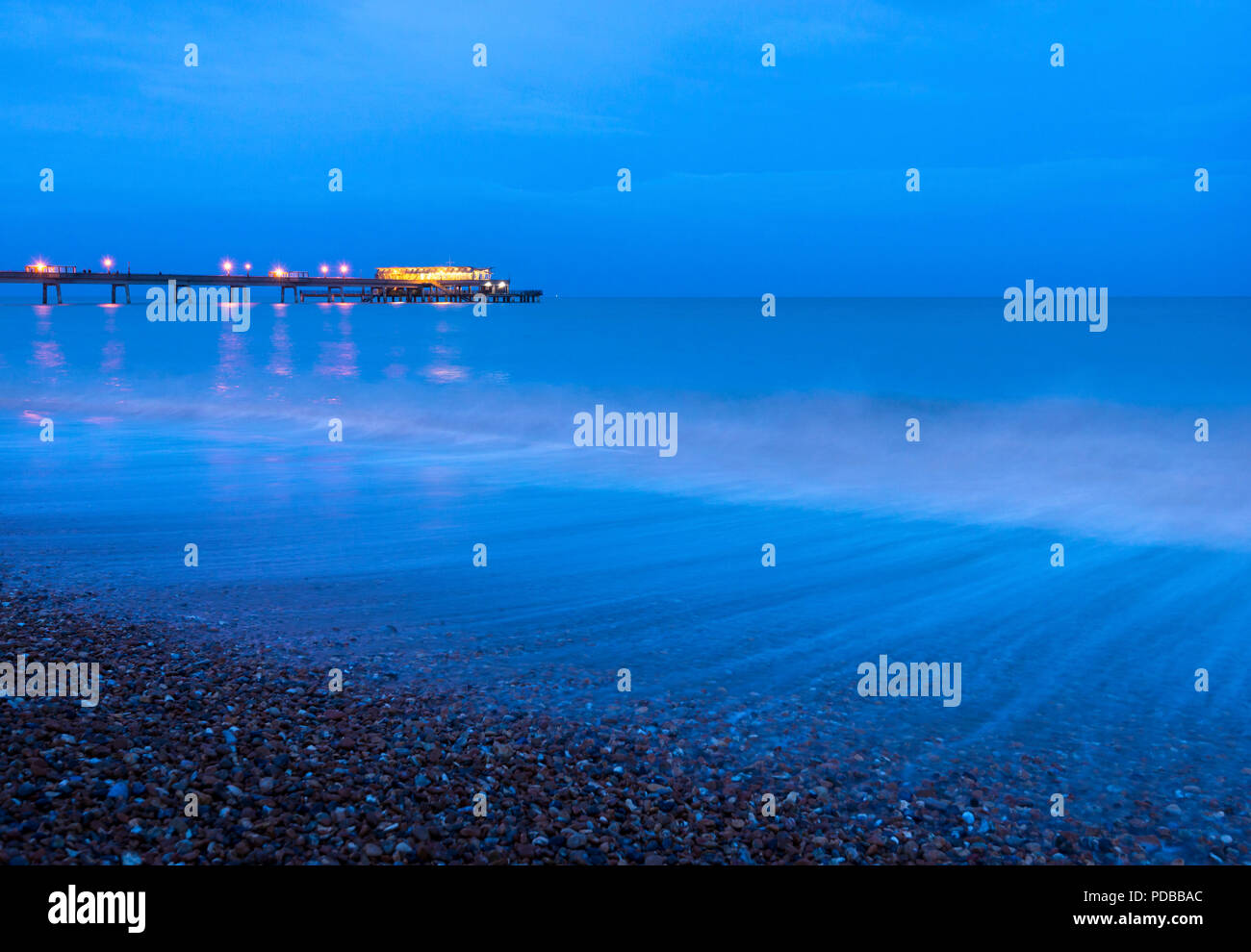 Onde che si infrangono lungo il lungomare di trattativa al tramonto con il Molo di trattativa illuminato in background. Foto Stock