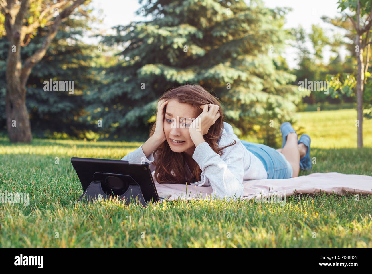 Ritratto di bianco giovane donna caucasica giacente nel parco su erba guardando digitale compressa. Una studentessa preoccupato sensazione ha sottolineato. Forte emozione, funny f Foto Stock