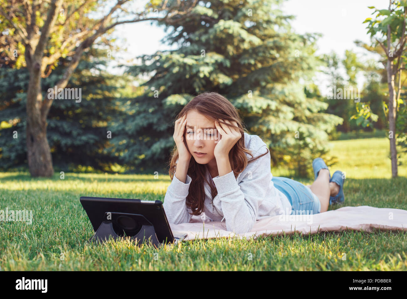 Ritratto di bianco giovane donna caucasica giacente nel parco su erba guardando digitale compressa. Una studentessa preoccupato sensazione ha sottolineato. Forte emozione, funny f Foto Stock