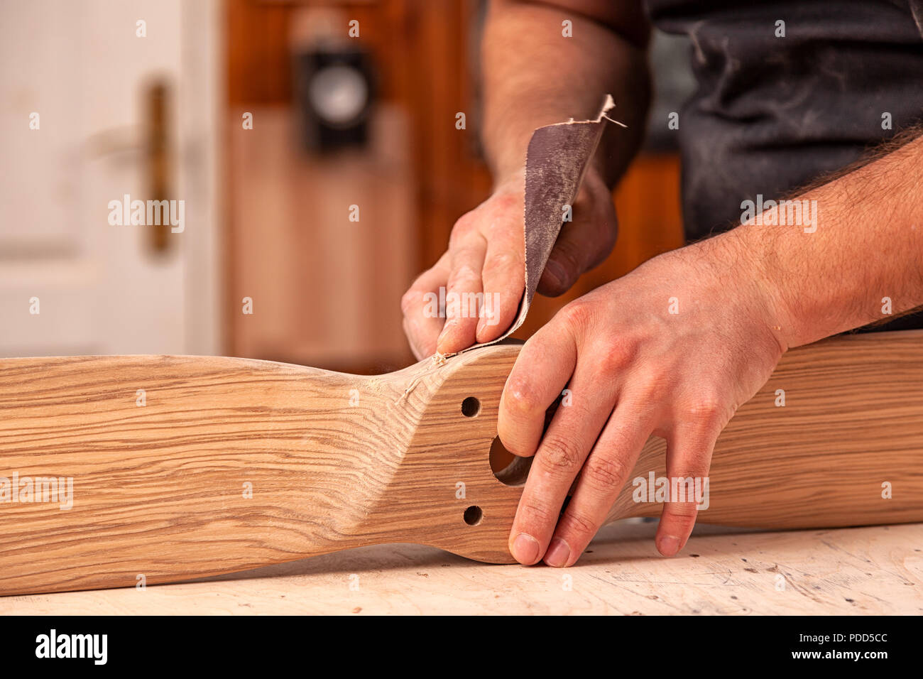 Un uomo di lavoro in un cappuccio e camicia lucida il blocco di legno con carta vetrata prima della verniciatura in officina, in background, strumenti e mach di perforazione Foto Stock