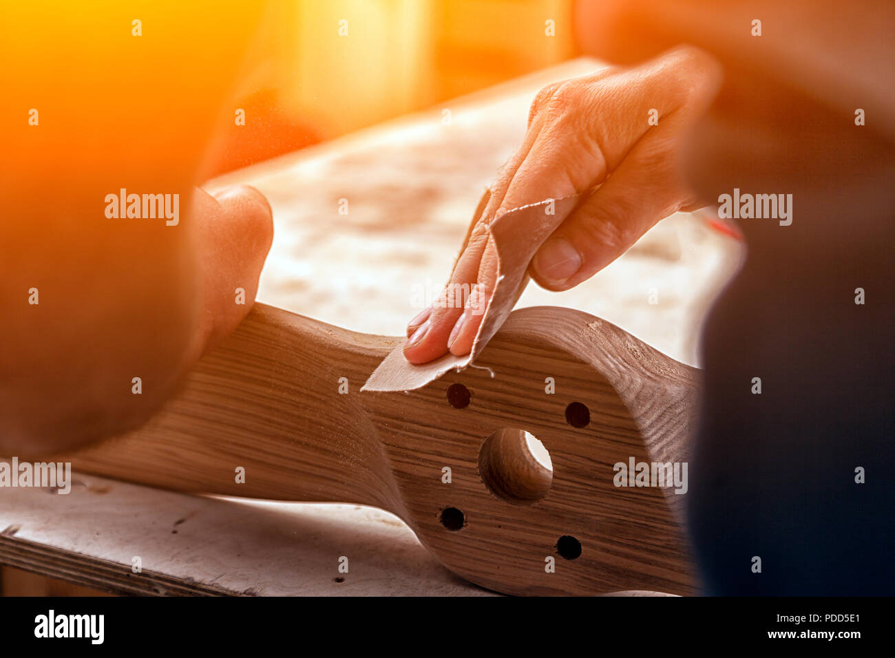 Un uomo di lavoro in un cappuccio e camicia lucida il blocco di legno con carta vetrata prima della verniciatura in officina, in background, strumenti e mach di perforazione Foto Stock