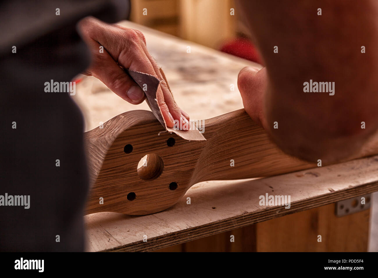 Un uomo di lavoro in un cappuccio e camicia lucida il blocco di legno con carta vetrata prima della verniciatura in officina, in background, strumenti e mach di perforazione Foto Stock