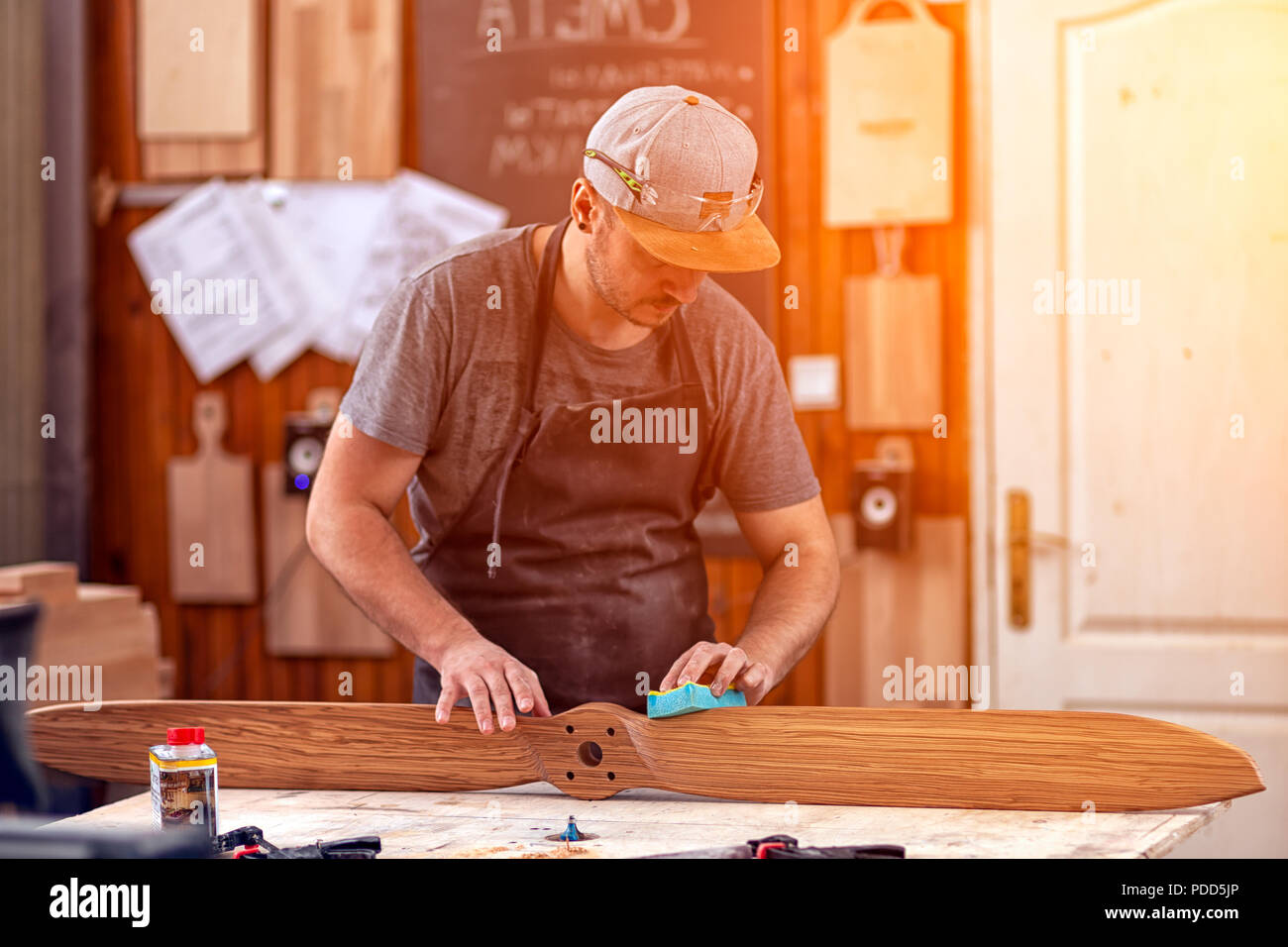Un uomo di lavoro in un cappuccio e camicia lucida il blocco di legno con carta vetrata prima della verniciatura in officina, in background, strumenti e mach di perforazione Foto Stock