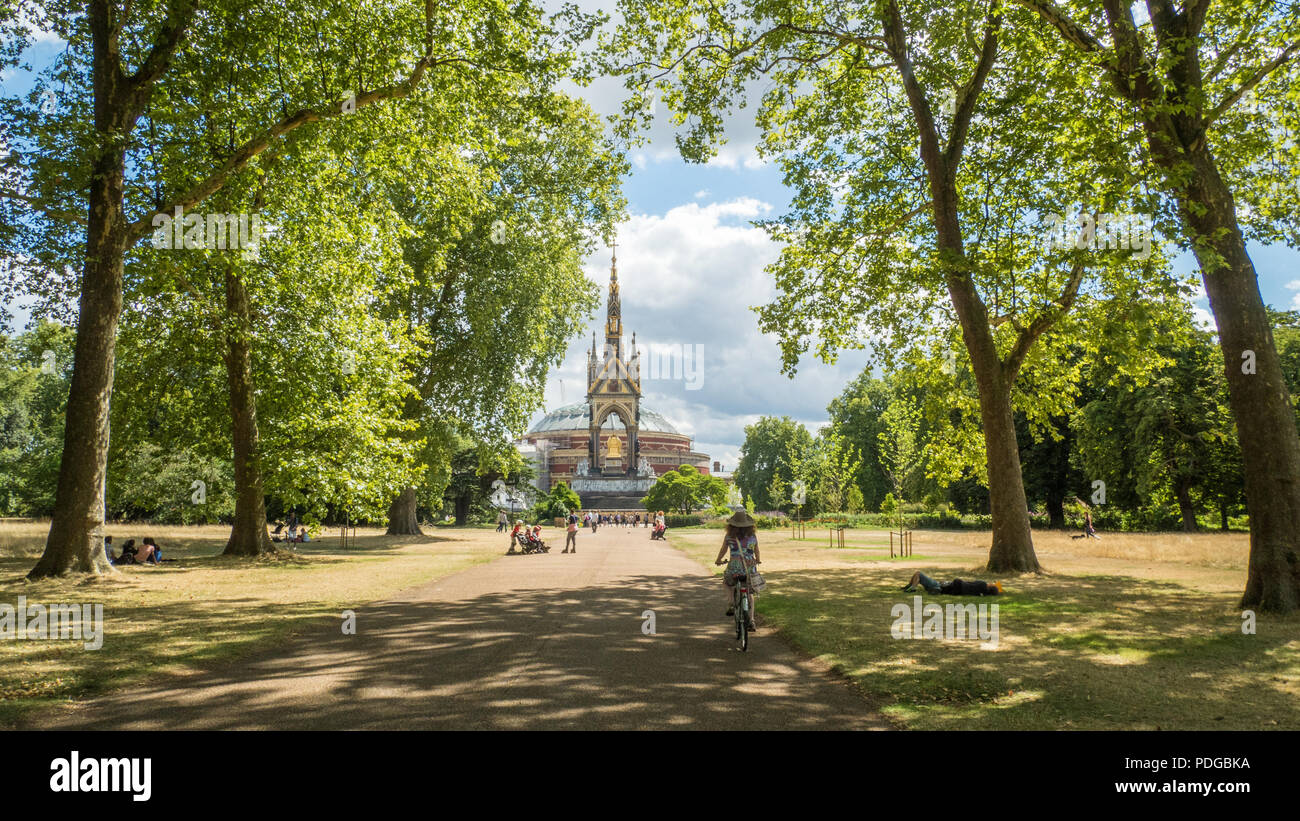 Lady pedalando vicino al monumento in stile gotico Albert Memorial a Kensington Gardens Royal Albert Concert Hall Behind, South Kensington, Londra. Foto Stock