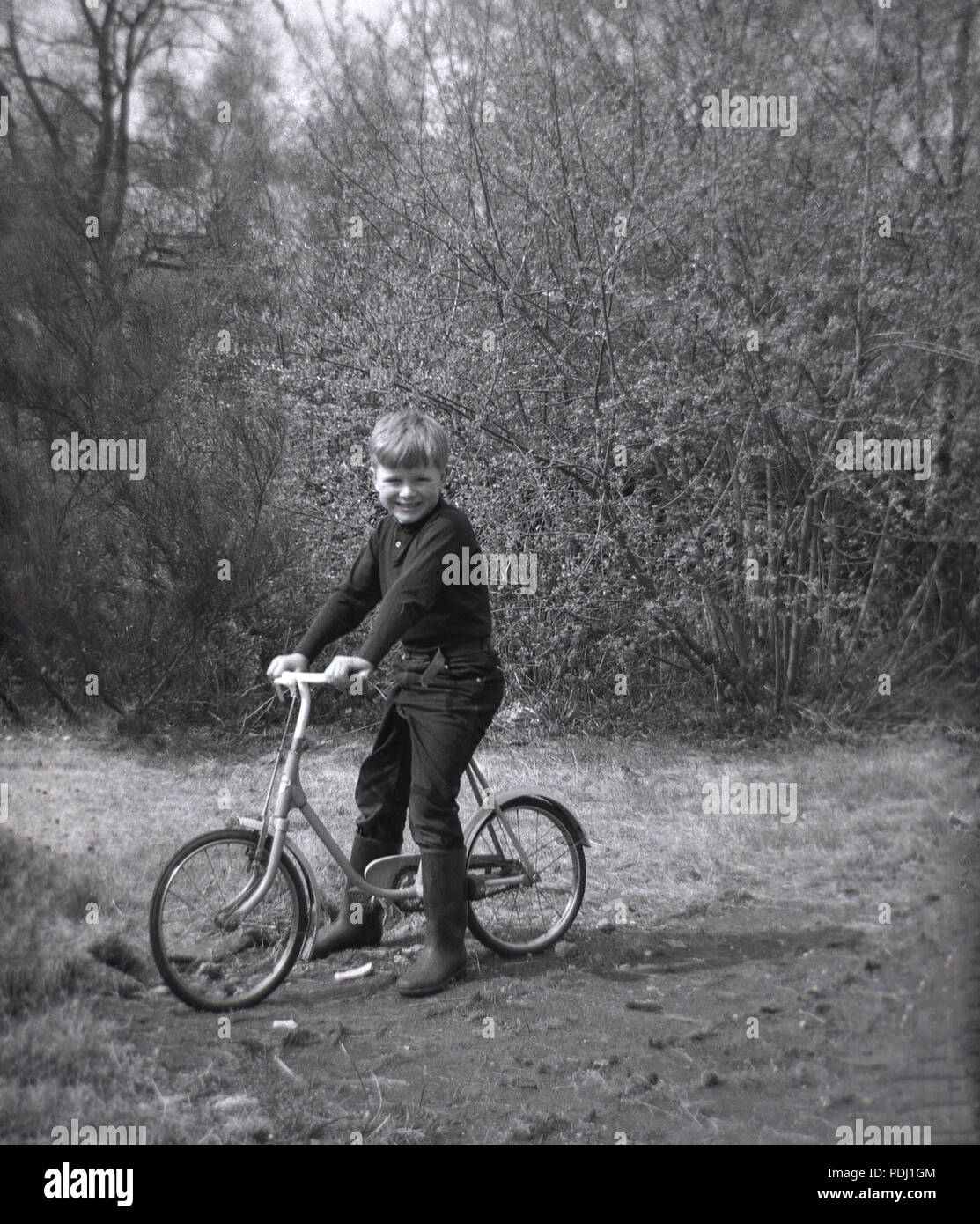 Anni sessanta, storico, ragazzo seduto su un piccolo a due ruote del bambino al di fuori di bicicletta su una pista da una foresta, Inghilterra, Regno Unito. Foto Stock