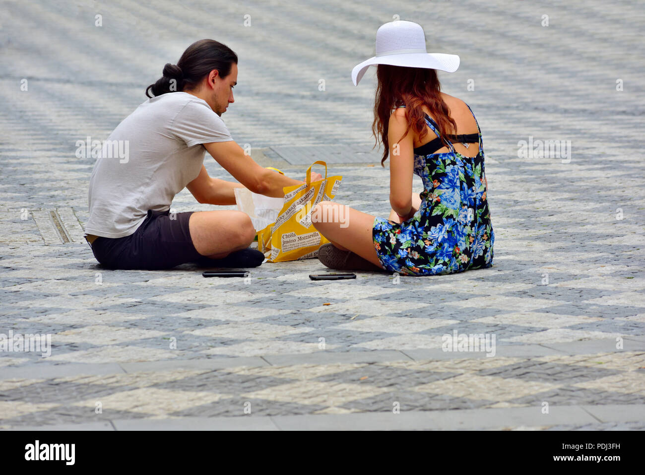 Coppia giovane seduto su cobblestone pavement in piazza snacking sul pranzo, godendo del sole Foto Stock