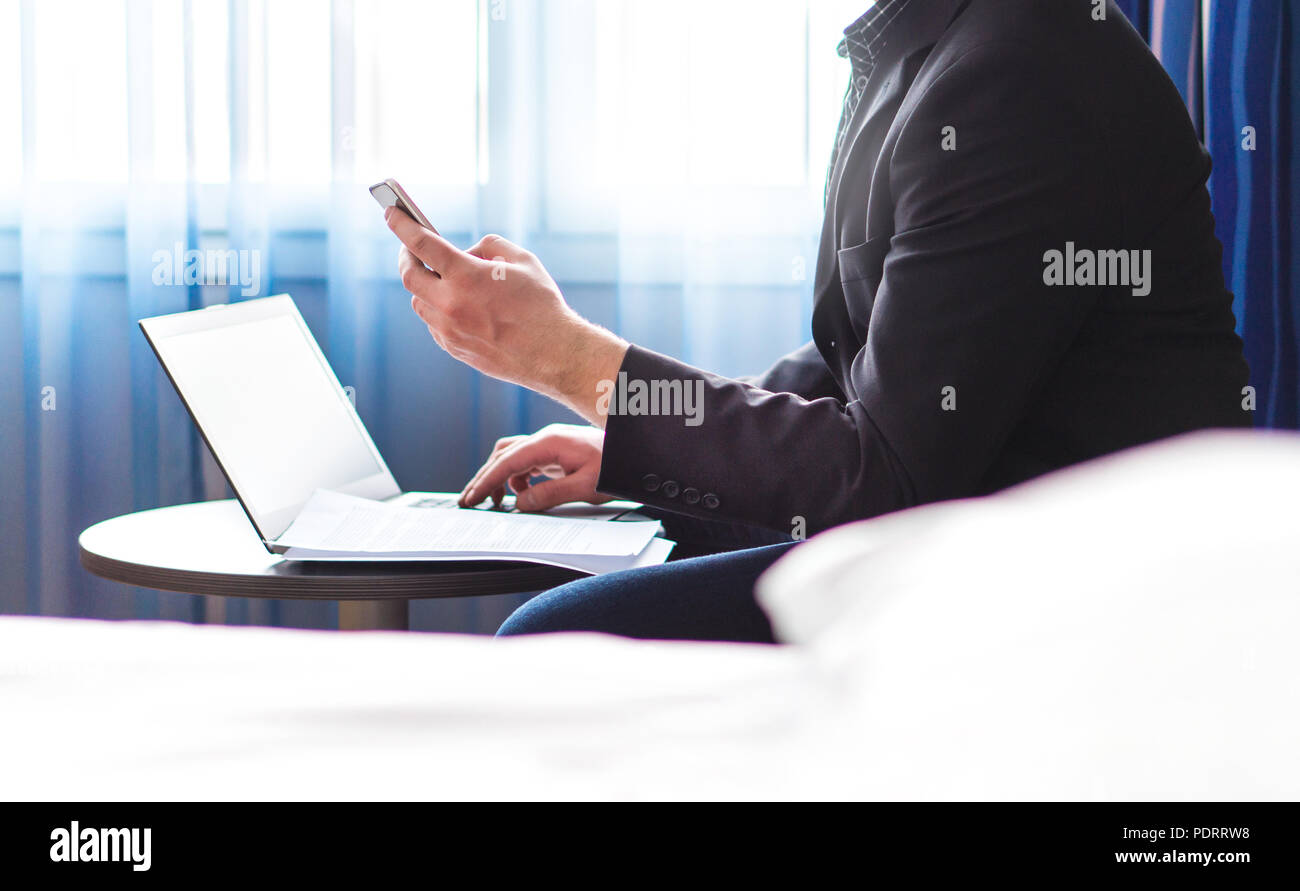 Business man in camera in hotel con lo smartphone e il computer portatile. Imprenditore la lettura dei messaggi di posta elettronica oppure utilizzando i social media. Il lavoro remoto durante il viaggio di affari. Foto Stock