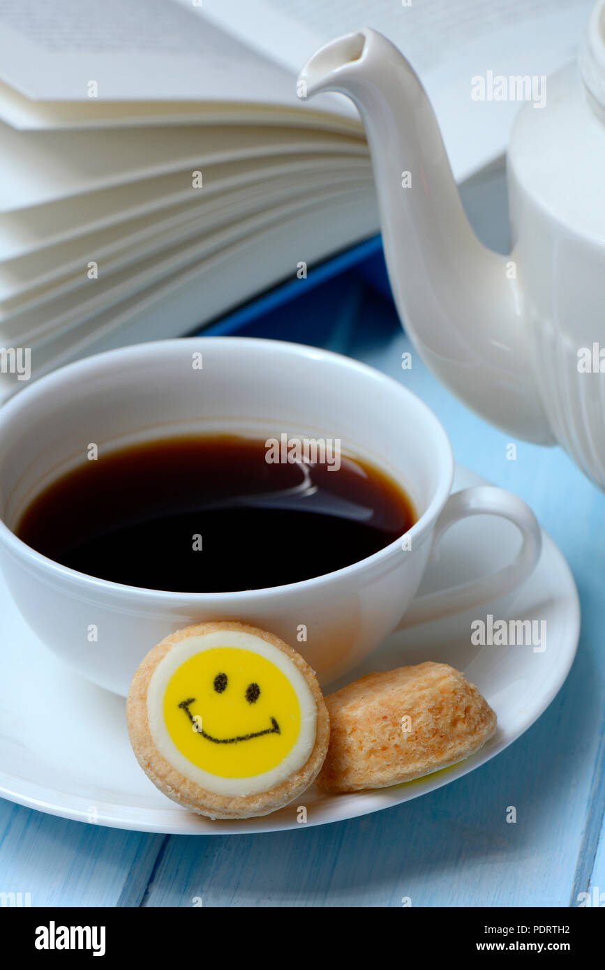Tasse Kaffee und Gebaeck mit Smiley, aufgeschlagenes Buch Foto Stock